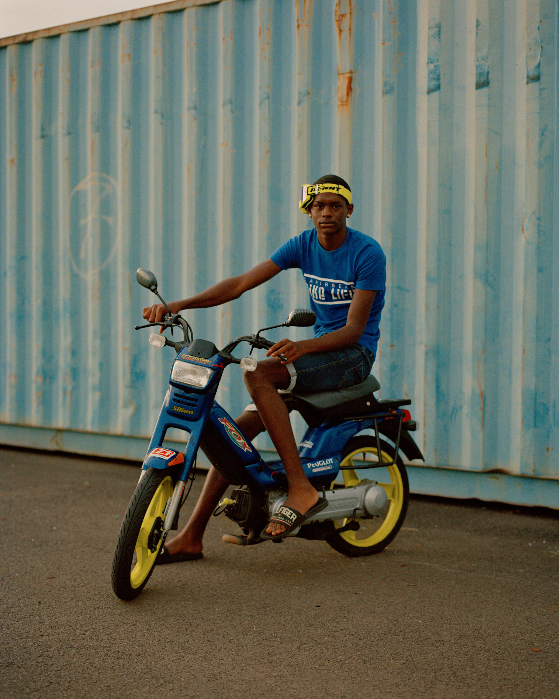 A young man sitting on a blue motorbike with yellow spokes. He wears a blue T-shirt that says 